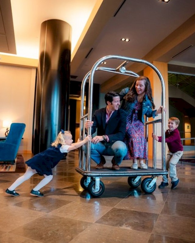 A family in a hotel lobby uses a luggage cart as a playful stroller, with kids pushing and pulling while parents smile at the scene.