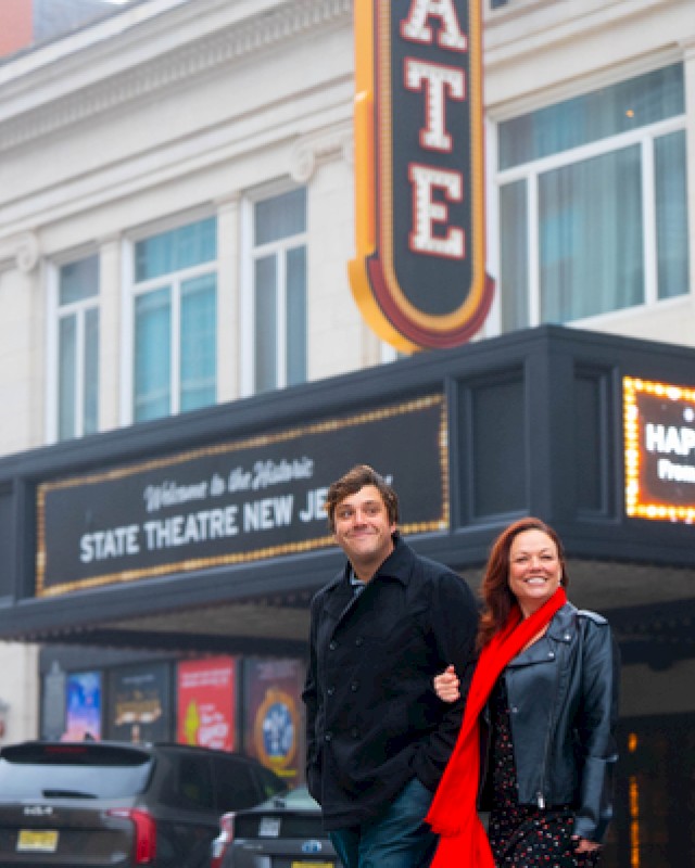 Two people walk arm-in-arm outside a theater marquee that reads “STATE THEATER NEW JERSEY,” with a bright red scarf and leather jacket.