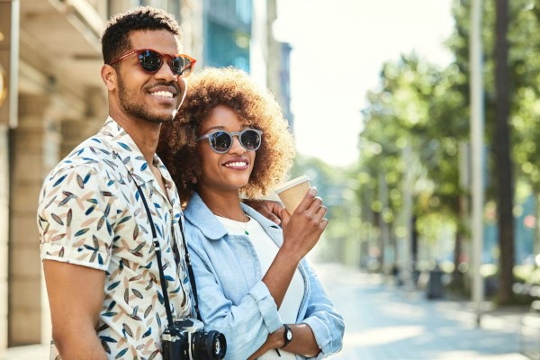 A smiling couple wearing sunglasses, one holding a camera and the other a coffee cup, walking outdoors on a sunny day.