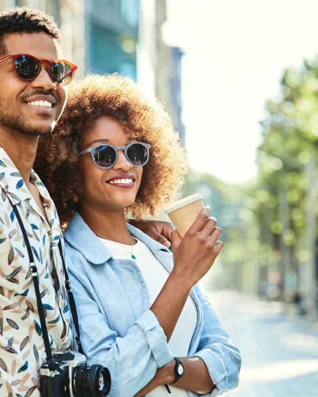 A smiling couple wearing sunglasses, one holding a camera and the other a coffee cup, walking outdoors on a sunny day.