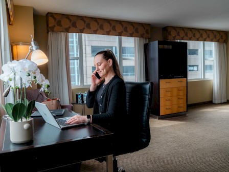 A woman is working on a laptop while talking on the phone in a hotel room with windows, curtains, and modern furniture.