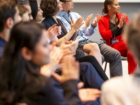 A diverse group of professionals and students sit in an auditorium, attentively watching and clapping during a presentation or event.