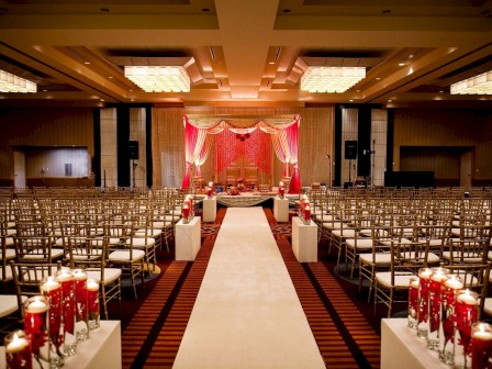 A grand wedding or ceremony hall with a central aisle, red draped stage, rows of chairs, and candlelit decor lining the aisle.
