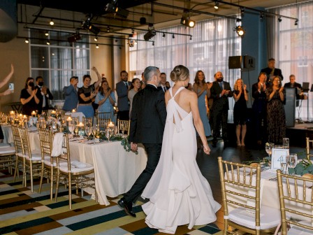 A couple in wedding attire walk hand in hand through a decorated reception hall with guests applauding; elegant tables, candles, and gold chairs abound.