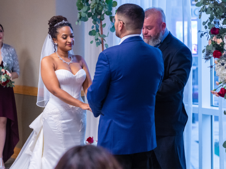 A bride in a white wedding dress exchanges vows with a groom in a blue suit, as an officiant stands nearby in a bright, floral ceremony room.