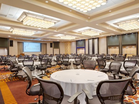 The image shows a conference room with round tables, chairs, and a presentation screen. The room has patterned carpet and ceiling lights.