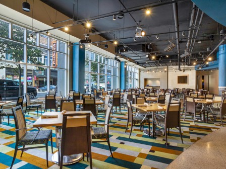 The image shows a spacious, modern dining area with many empty tables and chairs, colorful carpet, and large windows facing the street.