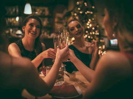 A group of women toasting with champagne in a warmly lit setting, possibly a celebration or gathering with a decorated tree in the background.