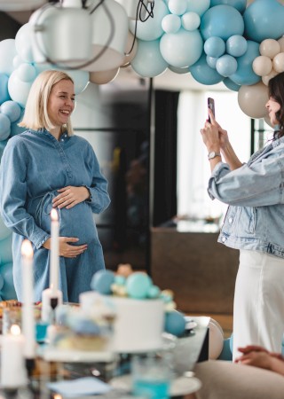 A pregnant woman is smiling at a party with blue and white balloons, while another woman takes a photo of her; celebration likely for a baby shower.