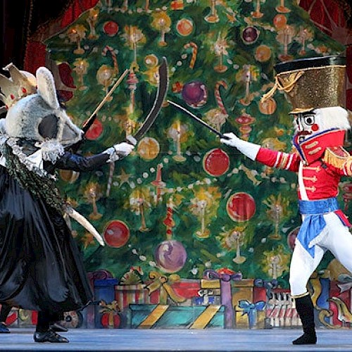 A costumed performance with a Nutcracker character fighting a mouse-like figure, set against a festive backdrop featuring a large Christmas tree.