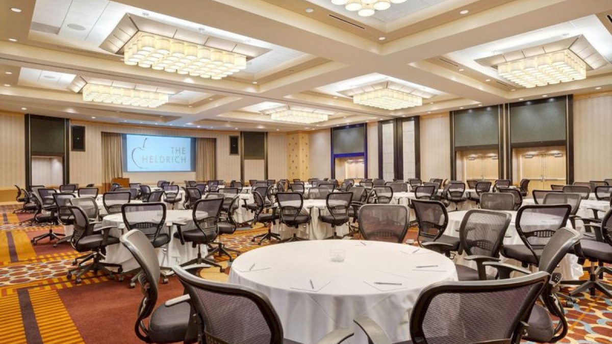 Photo of meeting room in cabaret or round table set up with black wheeled chairs, multiple windows and a white screen to project in the back of the meeting room