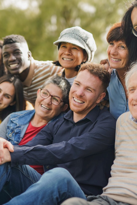 A diverse group of people are smiling and sitting outdoors, enjoying a sunny day together.