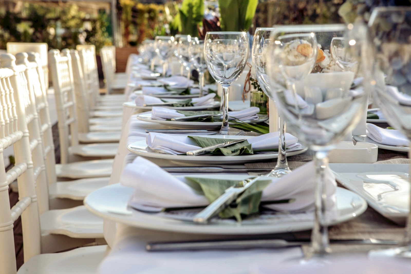 A beautifully set outdoor dining table prepared for a formal event or celebration with white plates, glasses, napkins, and green leaves.
