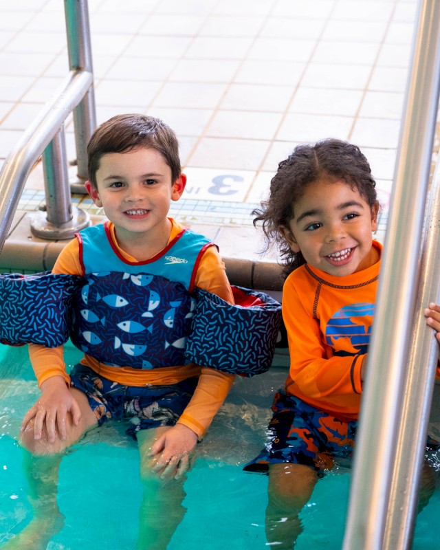 Two smiling kids sit in a pool, wearing orange and blue swimsuits with arm floaties, inside a metal pool step area.