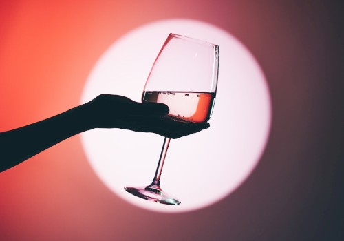 A hand holding a wine glass with a pink beverage, silhouetted against a large, out-of-focus circular light in the background.