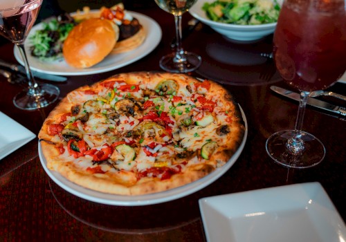 A table with a pizza topped with veggies, salad, burger, and drinks. There are also empty plates, ready for serving.
