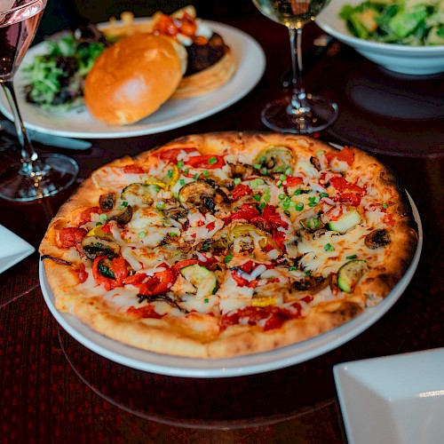 A table with a pizza topped with veggies, salad, burger, and drinks. There are also empty plates, ready for serving.