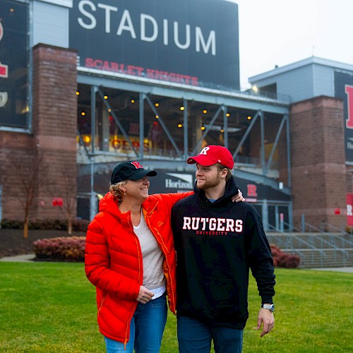 Two people wearing Rutgers gear stand in front of a stadium with 