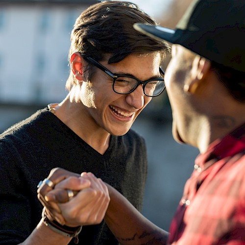 Two people are smiling and shaking hands outdoors, exuding a sense of friendship or camaraderie.