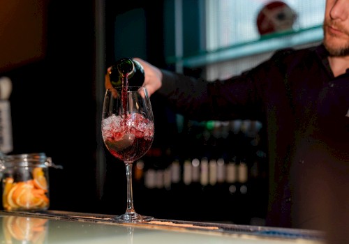 A person is pouring red wine into a glass on a bar counter with a jar of citrus slices nearby, under a dimly lit ambiance.