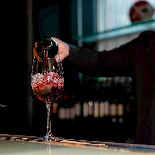 A person is pouring red wine into a glass on a bar counter with a jar of citrus slices nearby, under a dimly lit ambiance.