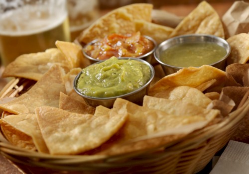 A basket of tortilla chips with guacamole, salsa, and green sauce, accompanied by a glass of beer on the side.