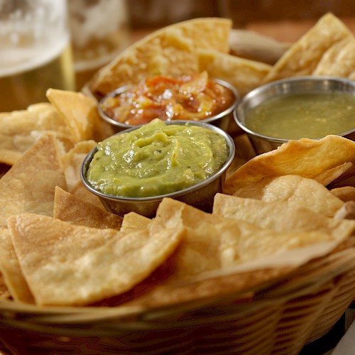 A basket of tortilla chips with guacamole, salsa, and green sauce, accompanied by a glass of beer on the side.