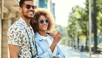 A smiling couple enjoys a sunny day outdoors, both wearing sunglasses; one holds a coffee cup and the other carries a camera.