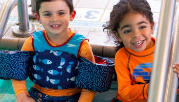Two children sitting at the edge of a pool, wearing bright swimwear and smiling, with feet in the water.