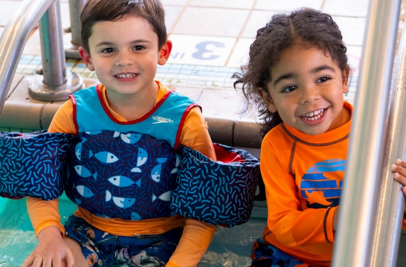 Two children sitting at the edge of a pool, wearing bright swimwear and smiling, with feet in the water.