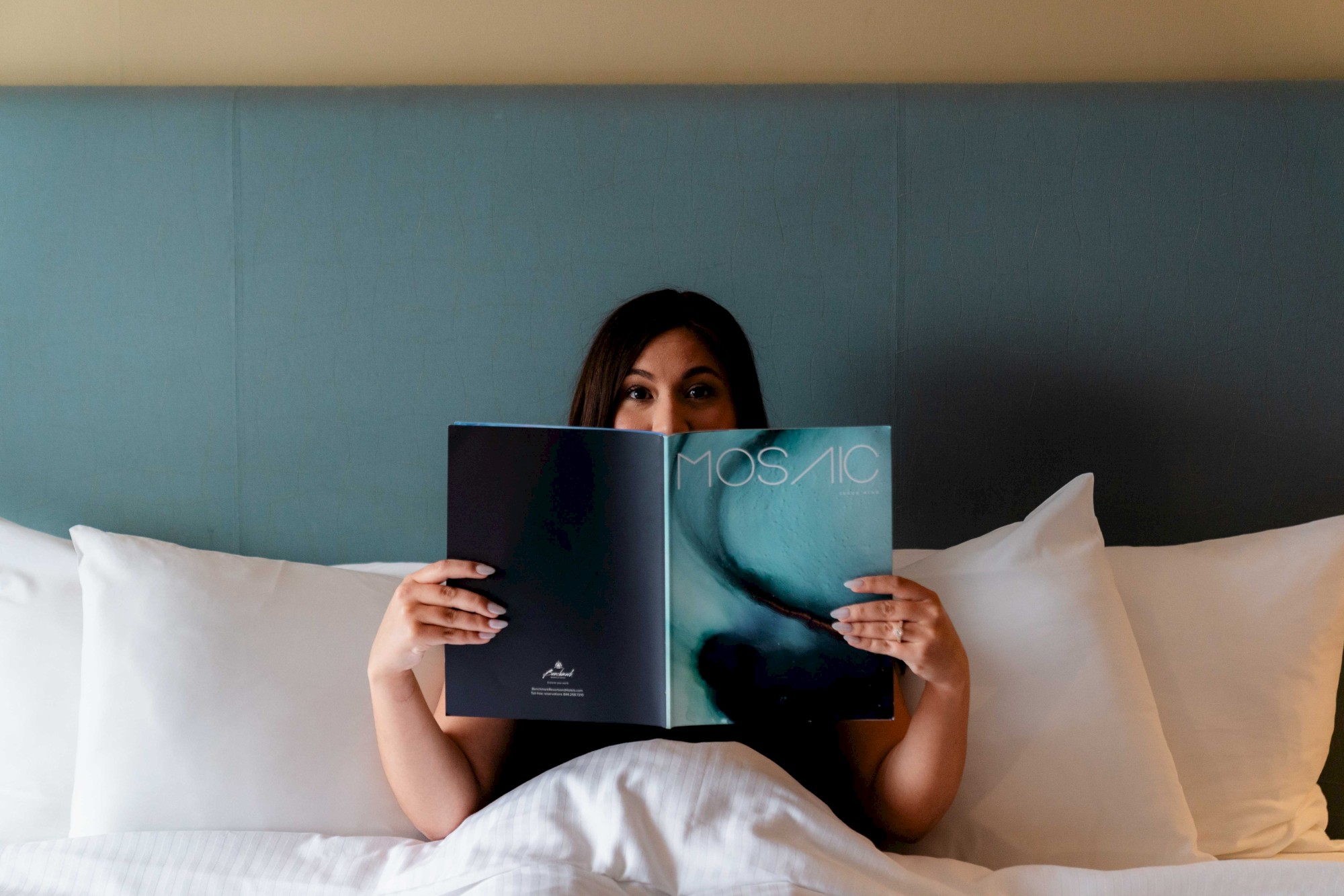 A person in bed with white pillows is holding up a magazine titled 