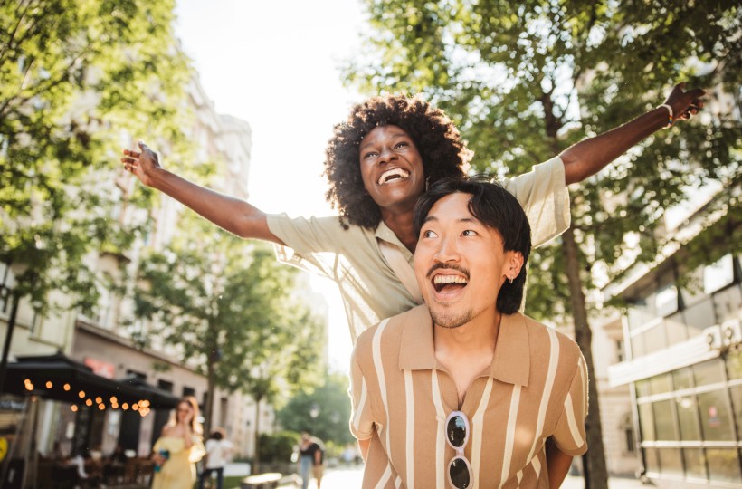 A joyful couple enjoys a piggyback ride on a sunny city street, surrounded by trees and buildings.