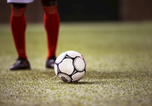 A soccer ball on a grassy field with a player in red socks and black shoes standing nearby, captured from a low angle.