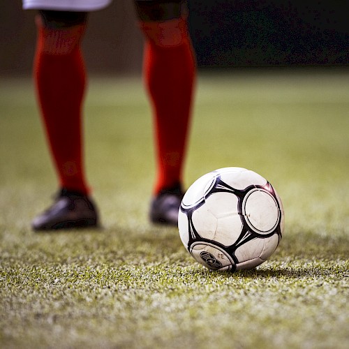 A soccer ball on a grassy field with a player in red socks and black shoes standing nearby, captured from a low angle.
