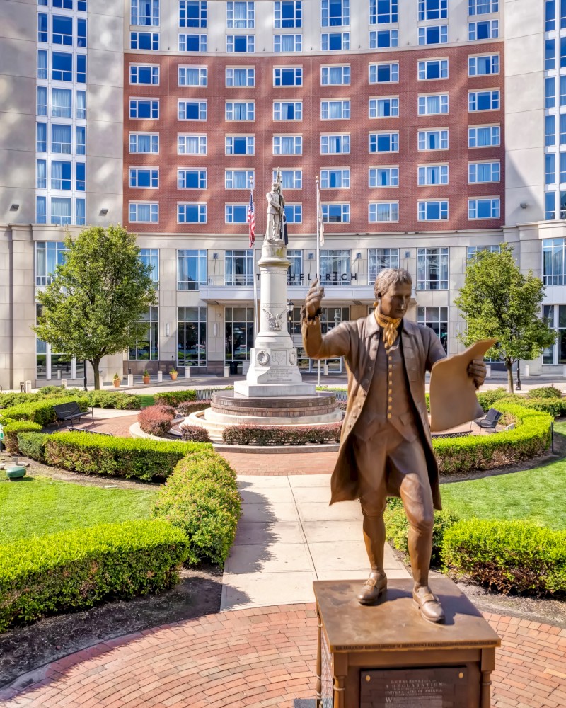 A statue of a man is in the foreground, set in a landscaped plaza with a large building in the background.