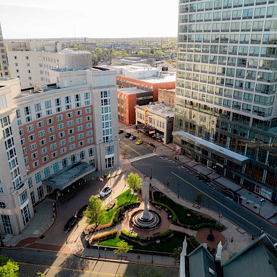 An aerial view of a cityscape featuring modern buildings, streets, and a roundabout with a central fountain surrounded by greenery.