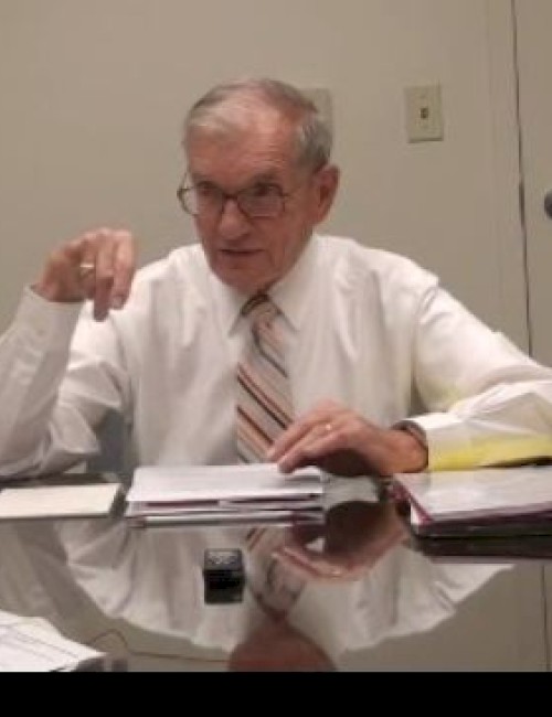 An elderly man in a shirt and tie sits at a table with documents and folders, appearing to engage in discussion or review.