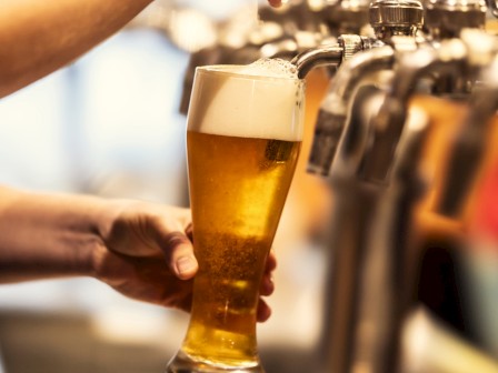 A person is pouring beer into a tall glass from a tap, with foam forming on top. Several beer taps are visible in the background.