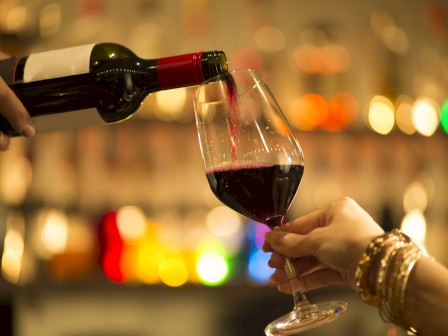 A person pours red wine into a glass held by another, with a blurred, colorful bokeh background in a cozy setting.