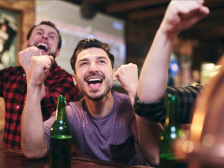 A group of excited people cheering at a bar, with drinks on the table and a TV screen in the background.