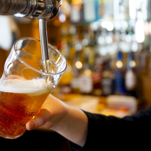 A person is pouring beer from a tap into a pint glass, with a blurred background of bar bottles and decor.