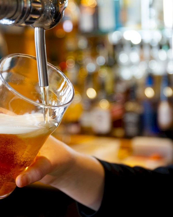 A person is pouring beer from a tap into a pint glass, with a blurred background of bar bottles and decor.