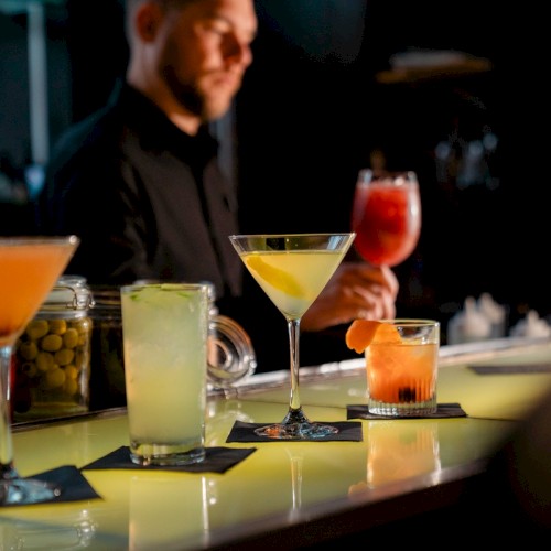 A bartender is preparing cocktails on a well-lit bar counter, with four different drinks displayed, including a martini and a fruity beverage.