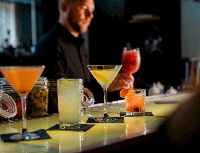 A bartender is preparing cocktails on a well-lit bar counter, with four different drinks displayed, including a martini and a fruity beverage.