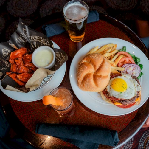A table with a burger topped with a fried egg, fries, wings with dip, a beer, and a cocktail.