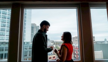 A couple stands by a large window in a high-rise, sharing a moment and possibly exchanging rings, with a cityscape view behind them.