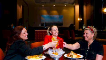 Three people are toasting with drinks at a table, enjoying food in a cozy restaurant setting.