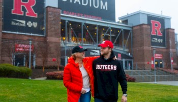 A man and woman, both wearing Rutgers attire, are walking in front of a stadium with large 