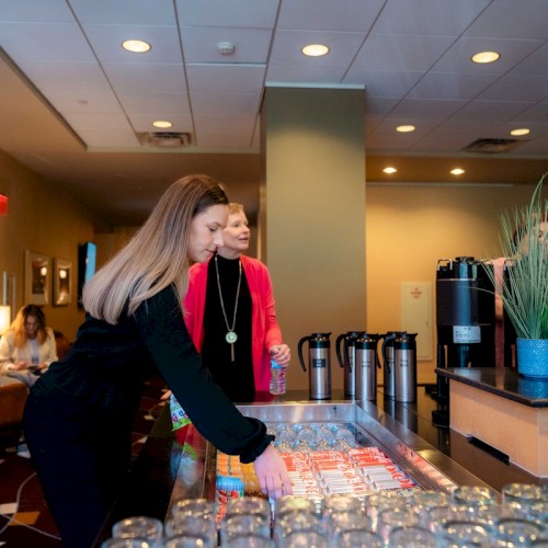 Two people are selecting drinks from a refreshment table with glass jars, plants, and coffee urns in a well-lit room.