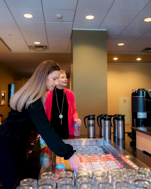 Two people are selecting drinks from a refreshment table with glass jars, plants, and coffee urns in a well-lit room.